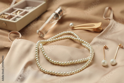 Stylish jewelry with pearls, luxury perfume and silk dress on beige fabric, closeup