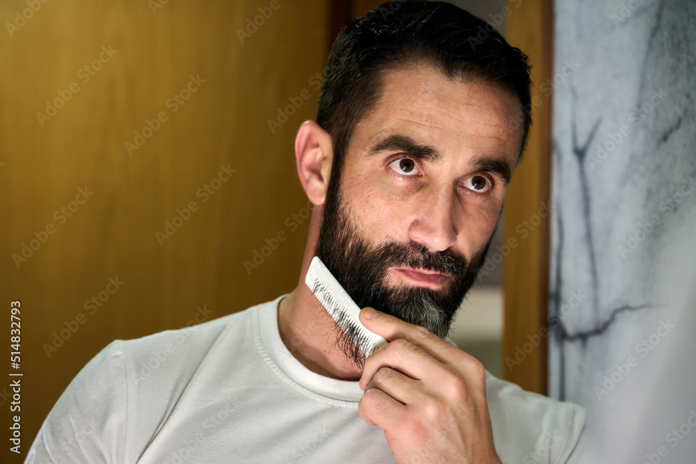 Young man grooming and combing his beard with comb in front of mirror