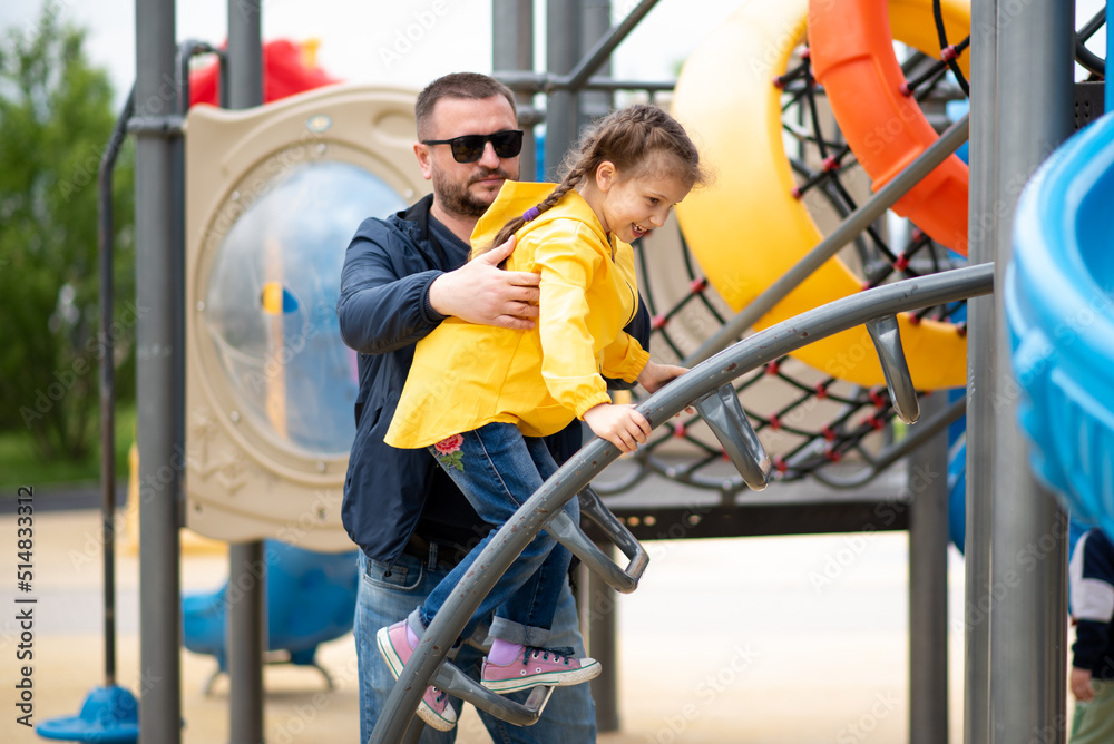 A girl 6-7 years old with her dad on the playground. She on the slide and has a fun. Dad helps the girl climb the stairs. Childhood. Time with family. Spring. City.