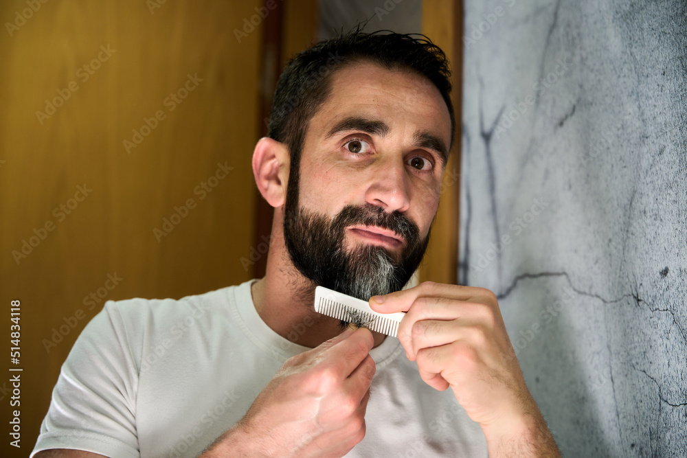 Young man grooming and combing his beard with comb in front of mirror Young man grooming and combing his beard with comb in front of mirror