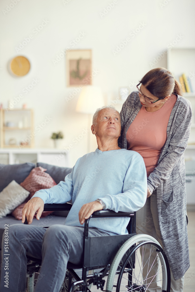 Serious senior woman in glasses pushing wheelchair with husband while taking care of him at home