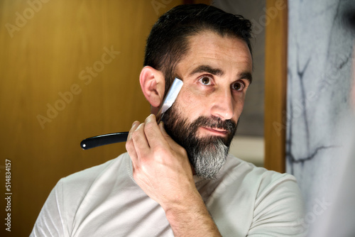 Young man with a beard shaving with a traditional knife in front of the mirror