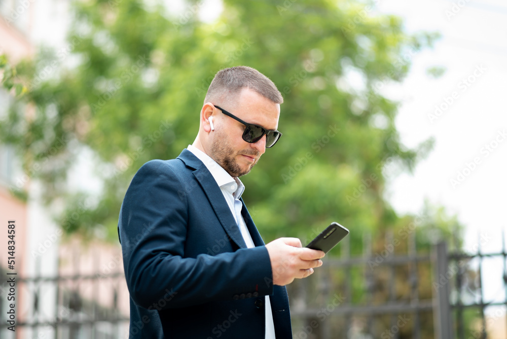 A business man in dark sunglasses with a phone in his hands stands outside the business building and talks on the phone. He is dressed in a light shirt and casual style jacket. A business man in dark sunglasses with a phone in his hands stands outside the business building and talks on the phone. He is dressed in a light shirt and casual style jacket.