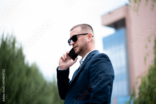 A business man in dark sunglasses with a phone in his hands stands outside the business building and talks on the phone. He is dressed in a light shirt and casual style jacket.