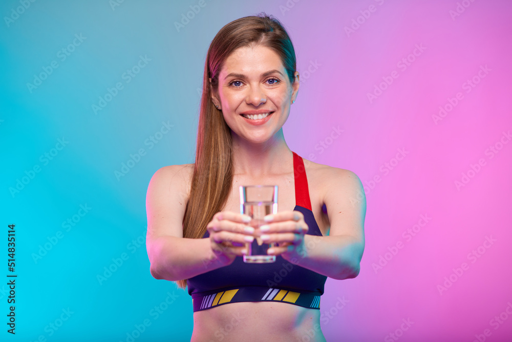 Smiling woman in sportswear holds water glass with two hands in front of her. Female fitness portrait isolated on neon multicolor background. Smiling woman in sportswear holds water glass with two hands in front of her. Female fitness portrait isolated on neon multicolor background.