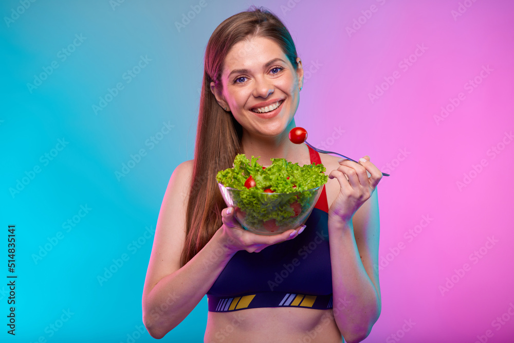 Smiling happy young woman in sportswear eating green salad in glass bowl. Female fitness portrait isolated on neon multicolor background. Smiling happy young woman in sportswear eating green salad in glass bowl. Female fitness portrait isolated on neon multicolor background.