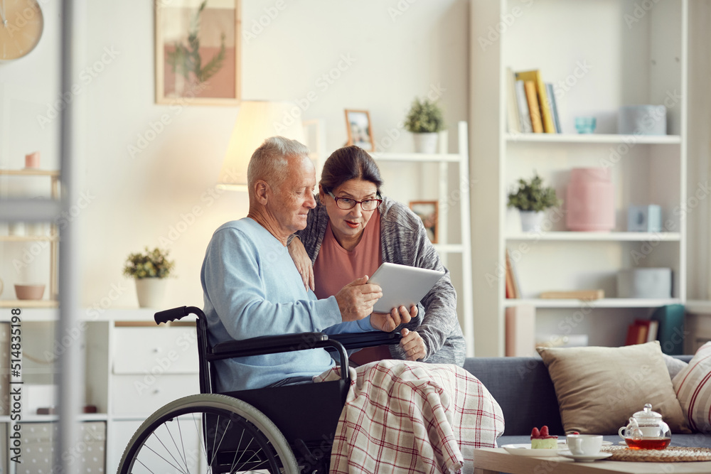 Senior man with legs covered with blanket sitting in wheelchair and showing new mobile app on tablet to surprised wife