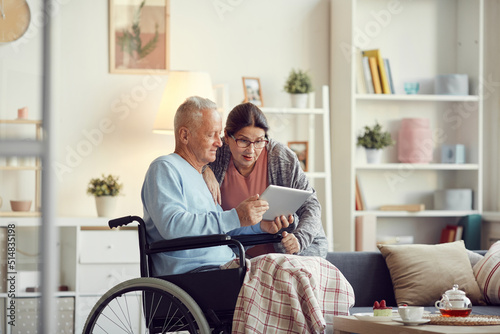 Senior man with legs covered with blanket sitting in wheelchair and showing new mobile app on tablet to surprised wife
