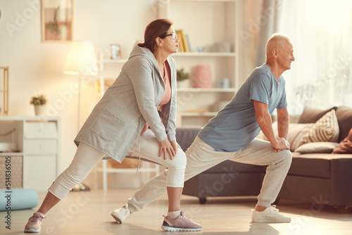 Energetic senior couple in sportswear leaning on knees while stretching legs at home training