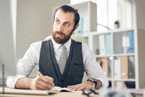 Serious thoughtful young bearded lawyer in gray waistcoat and white shirt sitting at table and making notes in planner while looking at computer monitor