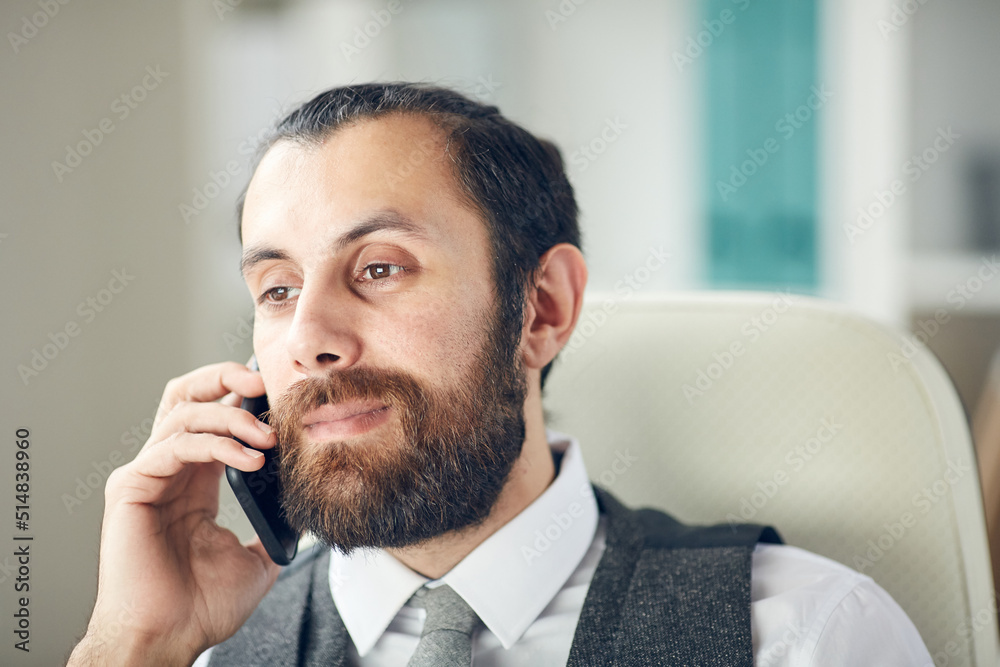 Close-up of serious busy male project manager with beard sitting on office chair and communicating by mobile phone Close-up of serious busy male project manager with beard sitting on office chair and communicating by mobile phone