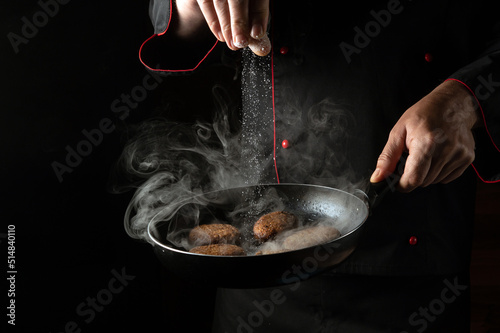 Fototapeta Naklejka Na Ścianę i Meble -  Adding salt to meat steaks in a hot pan in the hands of a chef. Cooking meat patties for a burger. Space for menu or recipe