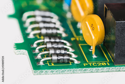 Semiconductor diodes and yellow resettable fuses in PCB detail on white background. Closeup of electronic components as diode or round polymeric PTC device - polyswitch on green printed circuit board. photo