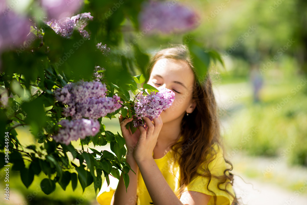 A teenage girl against the background of blooming lilacs enjoys spring and has fun. She has loose long wavy brown hair. She is dressed in a yellow t-shirt. Childhood. Beauty.