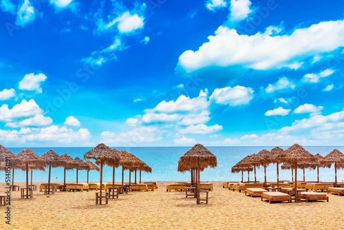 Sun loungers and umbrellas on the sandy beach by the sea and cloudy sky. Vacation background. Idyllic beach landscape on Mykonos island, Greece © Nikolay N. Antonov