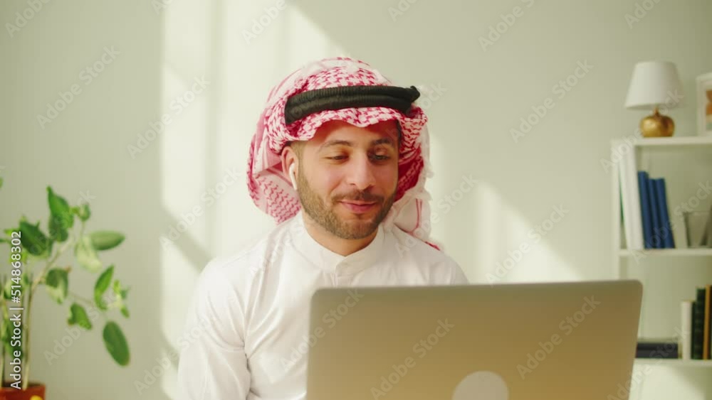 Middle eastern man calling on virtual conference, using earphones. Male person talking on video call on laptop. Wearing traditional Islamic clothes. Communicating with family and friends online. 