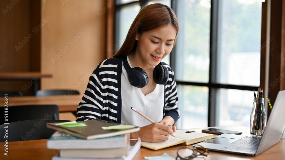Charming millennial Asian female college student with headphones doing math homework Charming millennial Asian female college student with headphones doing math homework