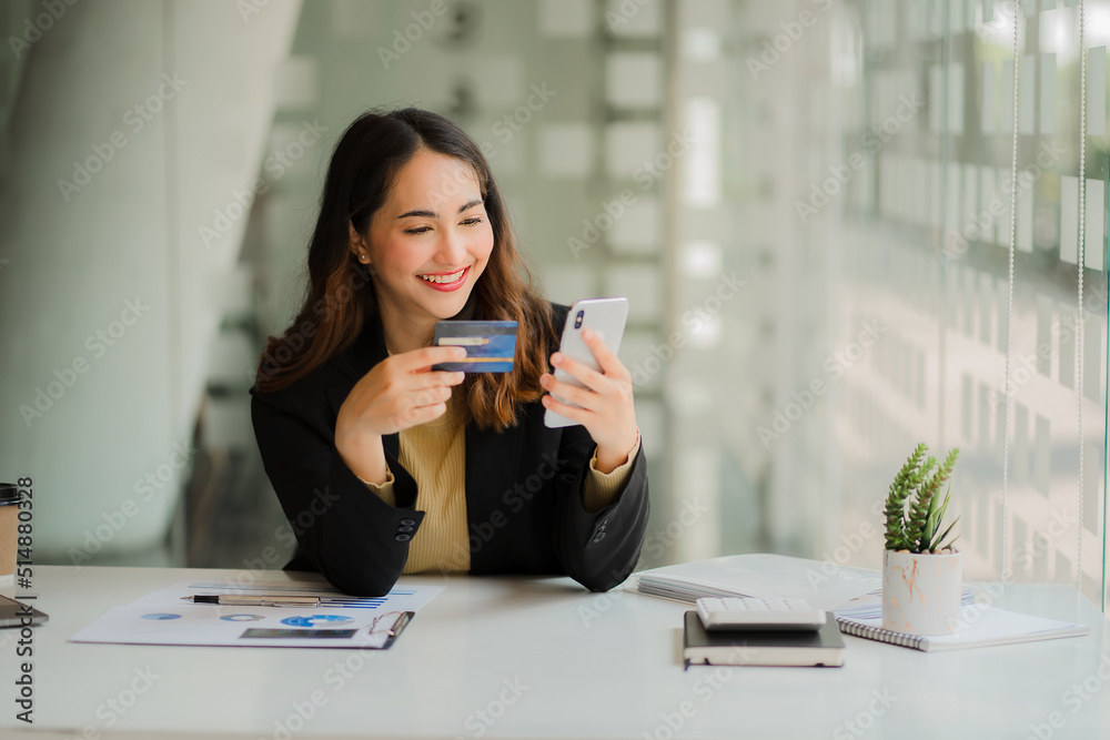 Asian business women use credit cards and mobile phones to pay and shop online with their laptop computers at their home desks. woman holding a debit card Internet Banking Payment Concept Asian business women use credit cards and mobile phones to pay and shop online with their laptop computers at their home desks. woman holding a debit card Internet Banking Payment Concept