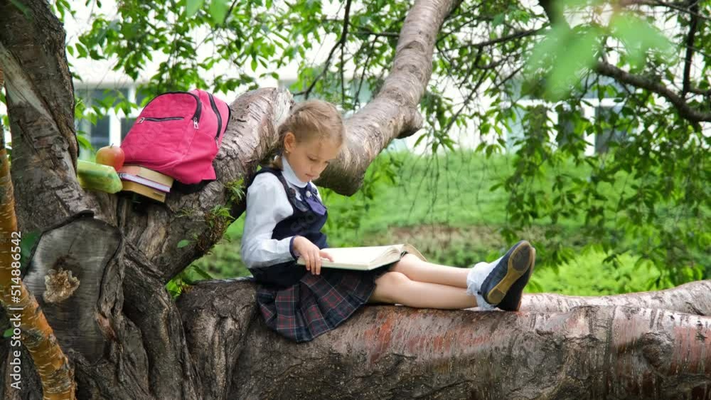 Little girl in school uniform relaxing on a tree branch and reading a book in spring or summer day. Literature, education and back to school concept Little girl in school uniform relaxing on a tree branch and reading a book in spring or summer day. Literature, education and back to school concept