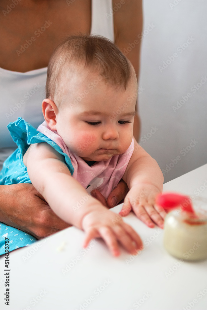 Child six month old with mother on a kitchen at a table with jar with vegetable puree and spoon. Baby girl tries to eat herself. Child six month old with mother on a kitchen at a table with jar with vegetable puree and spoon. Baby girl tries to eat herself.