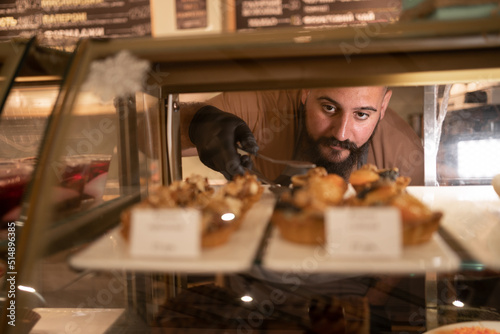 Indian man choosing variation of delicious cake in bakery showcase fridge at cafe. Male customer buying sweet cake at coffee shop. Small business restaurant food