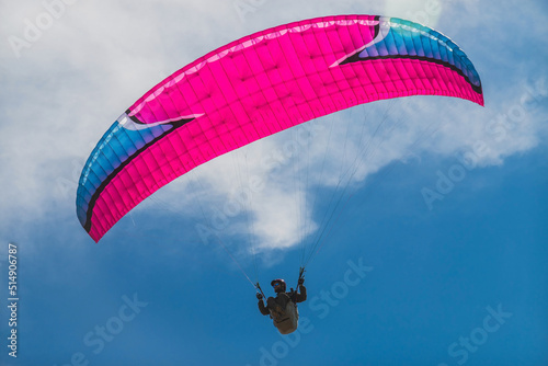A close-up paraglider pilot soars in the blue sky on a pink paraglider.