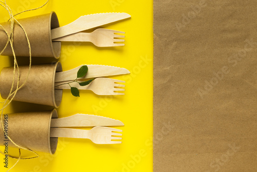 Paper-craft cups, wooden forks and knives on yellow. Eco-friendly disposable kitchenware utensils. Picnic kit. Ecology, zero waste concept. Environmental protection.Flat lay, copy space.