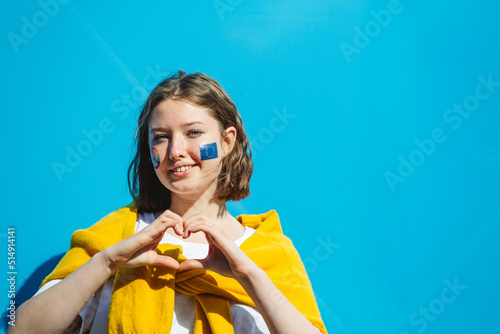 Smiling teenage girl with peace symbol and European Union paint on cheeks gesturing heart shape in front of blue wall