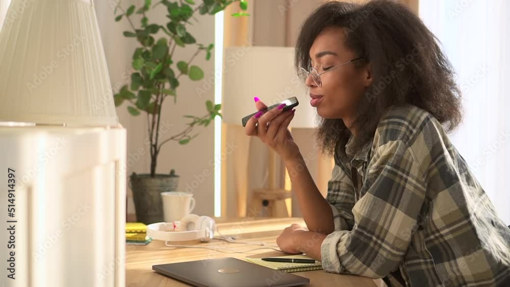 Young woman is holding phone and chatting online while sitting at table at home spbd. Close view of beautiful American African female holds smartphone in her hand and talks with smile, sits at desk Young woman is holding phone and chatting online while sitting at table at home spbd. Close view of beautiful American African female holds smartphone in her hand and talks with smile, sits at desk