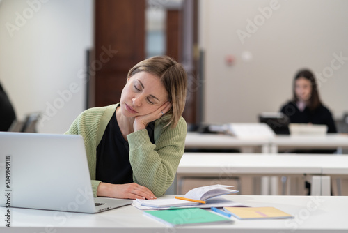 Tired female mature student falling asleep during boring online lesson, sleepy bored middle-aged woman getting second education sitting at desk with laptop in university library, selective focus