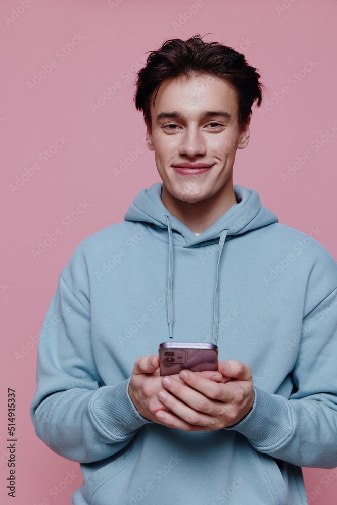 a handsome, attractive man stands on a pink background in a light blue hoodie and holding a smartphone in his hands looks at the camera with a friendly smile a handsome, attractive man stands on a pink background in a light blue hoodie and holding a smartphone in his hands looks at the camera with a friendly smile