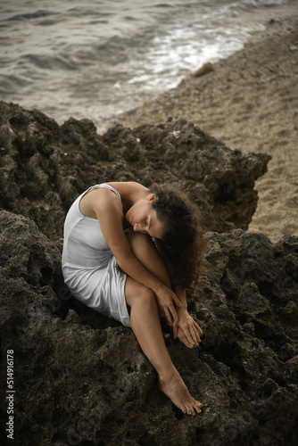A sadness woman is sitting on the rock. A curly-haired female in depression. Pensive brunette girl in white dress crying and worry at the sea. Dark photography