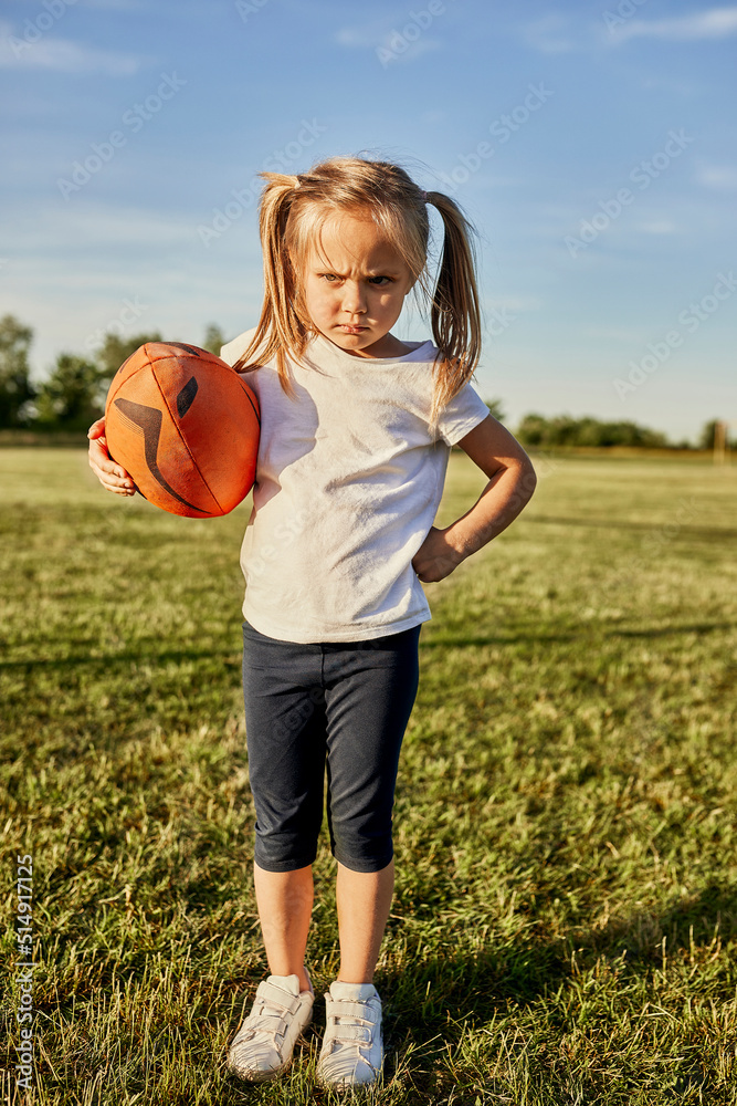 Angry blond girl with rugby ball standing at sports field on sunny day Angry blond girl with rugby ball standing at sports field on sunny day