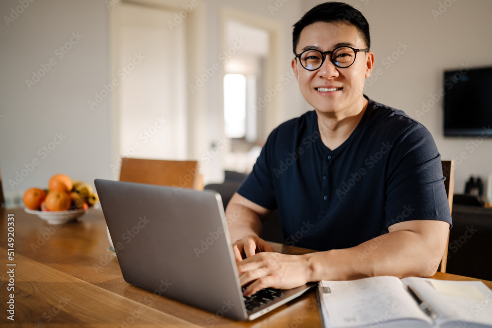 Brunette adult asian man smiling and using laptop computer at home