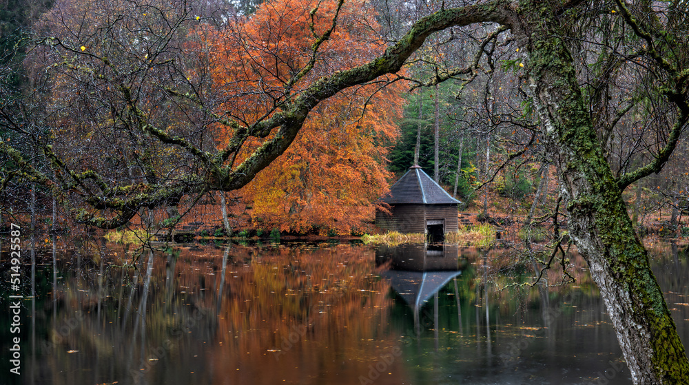 A moody, colourful autumn woodland forest over a calm reflection on Loch Dunmore near Pitlochry in Perthshire, Scotland.