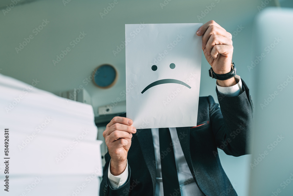 Unhappy businessman holding paper with frowning emoticon in office interior - obrazy, fototapety, plakaty 