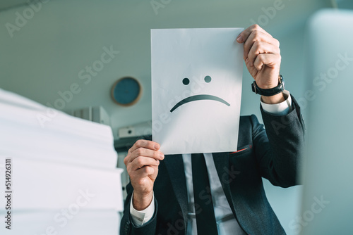 Fototapeta Unhappy businessman holding paper with frowning emoticon in office interior