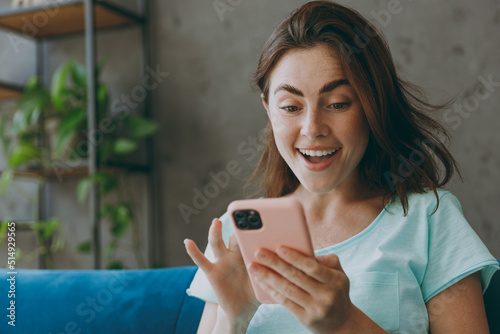 Young happy excited amzed caucasian woman she 20s in casual clothes mint t-shirt hold in hand use mobile cell phone sit on blue sofa indoor rest at home in own room apartment People lifestyle concept photo