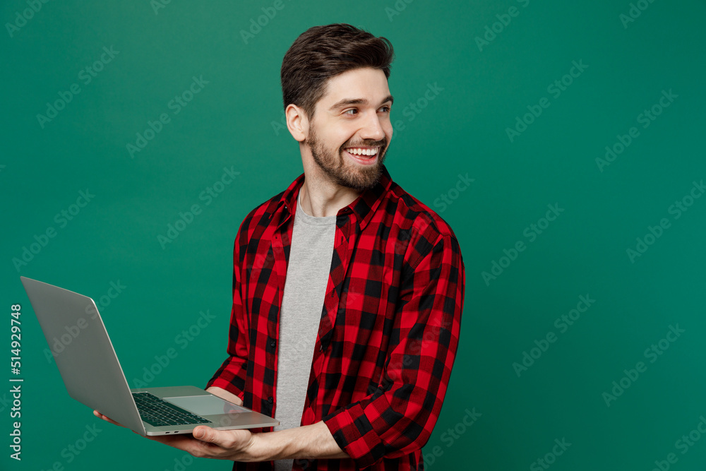 Young happy smiling cheerful fun cool man he 20s wear red shirt grey t-shirt hold use work on laptop pc computer look aside on workspace area isolated on plain dark green background studio portrait