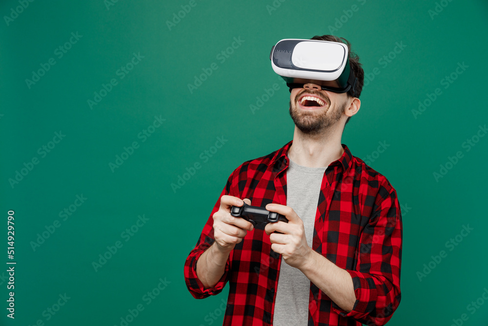 Young happy smiling man he 20s wearing red shirt grey t-shirt hold in hand play pc game with joystick console watching in vr headset pc gadget isolated on plain dark green background studio portrait Young happy smiling man he 20s wearing red shirt grey t-shirt hold in hand play pc game with joystick console watching in vr headset pc gadget isolated on plain dark green background studio portrait