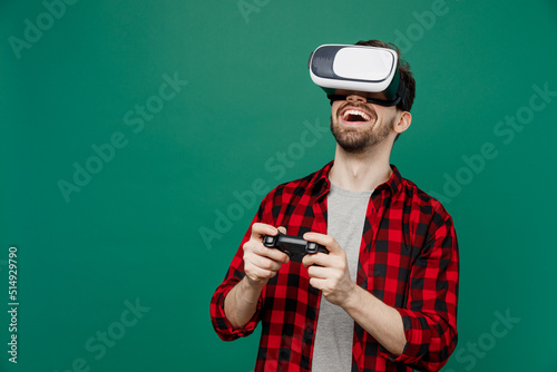 Young happy smiling man he 20s wearing red shirt grey t-shirt hold in hand play pc game with joystick console watching in vr headset pc gadget isolated on plain dark green background studio portrait