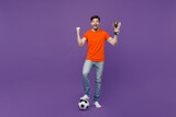 Full body young fan fun man he wear orange t-shirt cheer up support football sport team hold soccer ball champion cup do winner gesture watch tv live stream isolated on plain dark purple background.