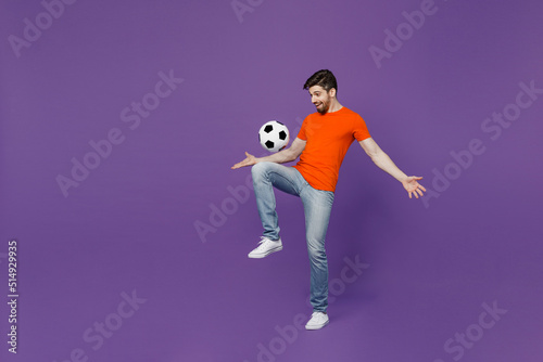 Full body young cheerful excited fun fan man he 20s wear orange t-shirt cheer up support football sport team juggling soccer ball on knee watch tv live stream isolated on plain dark purple background.