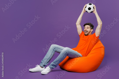 Full body excited young fan man he wear orange t-shirt cheer up support football sport team hold soccer ball above head watch tv live stream sit in bag chair isolated on plain dark purple background.