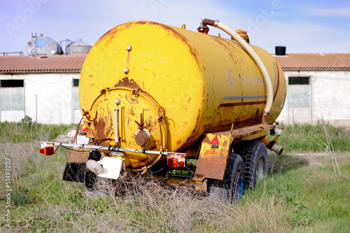 Fotografie A slurry tanker trailer, in disuse and in poor condition, is abandoned in the field next to a pig barn and is being engulfed by weeds and nature