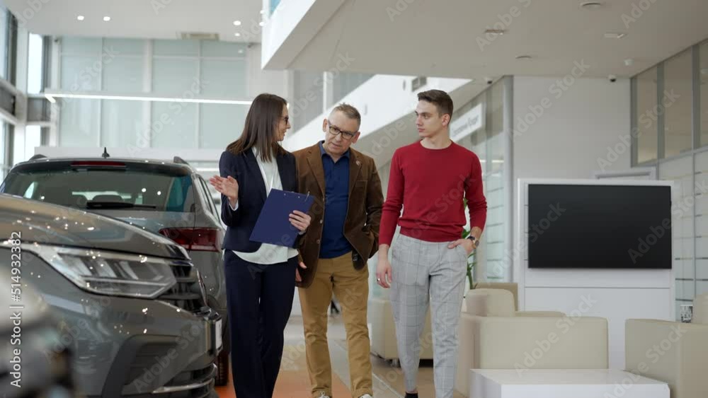 Two family people are talking to female agent and walking in car dealership office spbd. 4k Elderly father, young son walk in sales room and talk with woman consultant, study car model and make