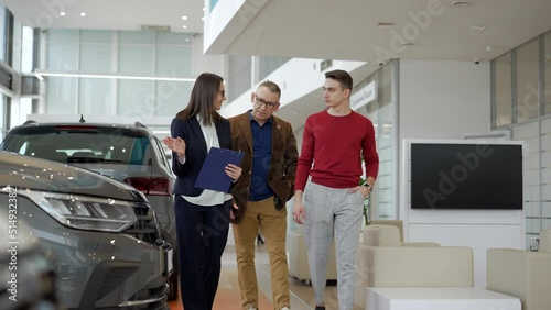 Two family people are talking to female agent and walking in car dealership office spbd. 4k Elderly father, young son walk in sales room and talk with woman consultant, study car model and make