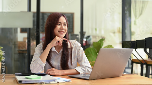 Happy female office worker sitting front of laptop and looking out of window, dreaming about future new successful projects