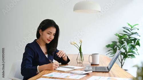 Happy young woman working with financial document and using laptop on wooden office desk