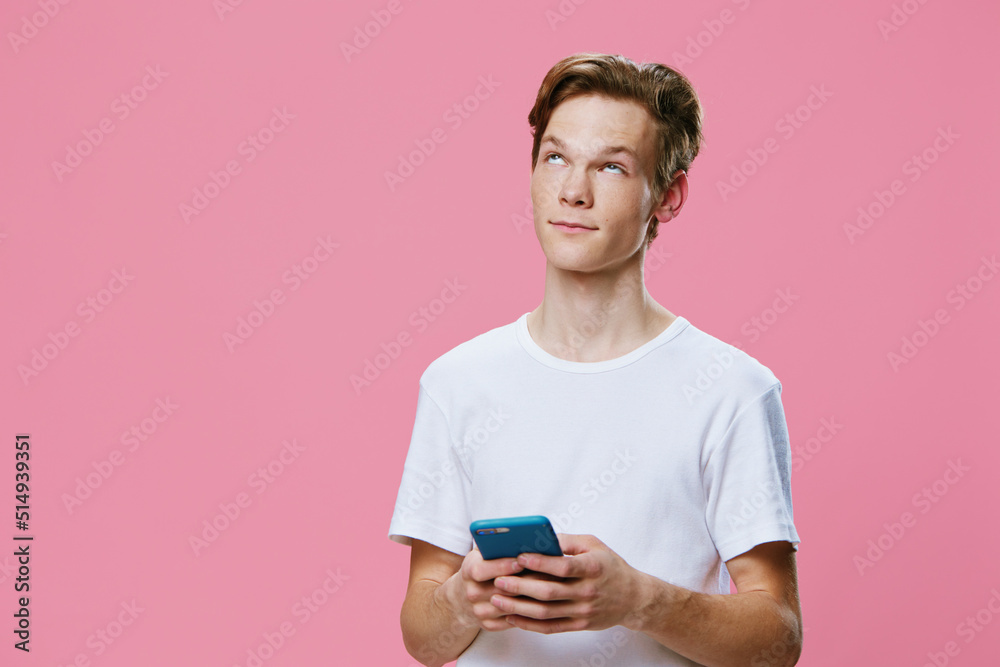 a handsome guy in a white t-shirt looks up thoughtfully while standing with a phone in his hands on a pink background a handsome guy in a white t-shirt looks up thoughtfully while standing with a phone in his hands on a pink background